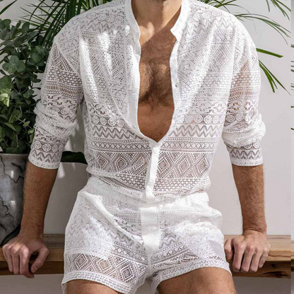 Mann in weißem, transparentem Spitzenhemd und Shorts, sitzt vor Pflanzen.