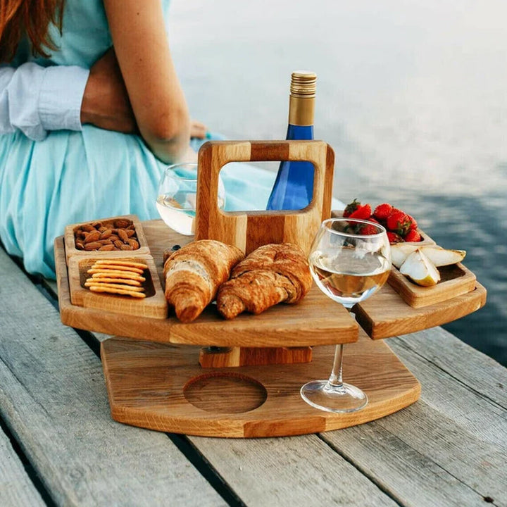 Wooden wine table on a dock with croissants, almonds, crackers, pear slices and strawberries, a person seated behind 3