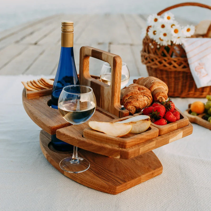 Wooden table on a white blanket with croissants, strawberries, pear slices, crackers, a wine bottle and glass, beside a 8