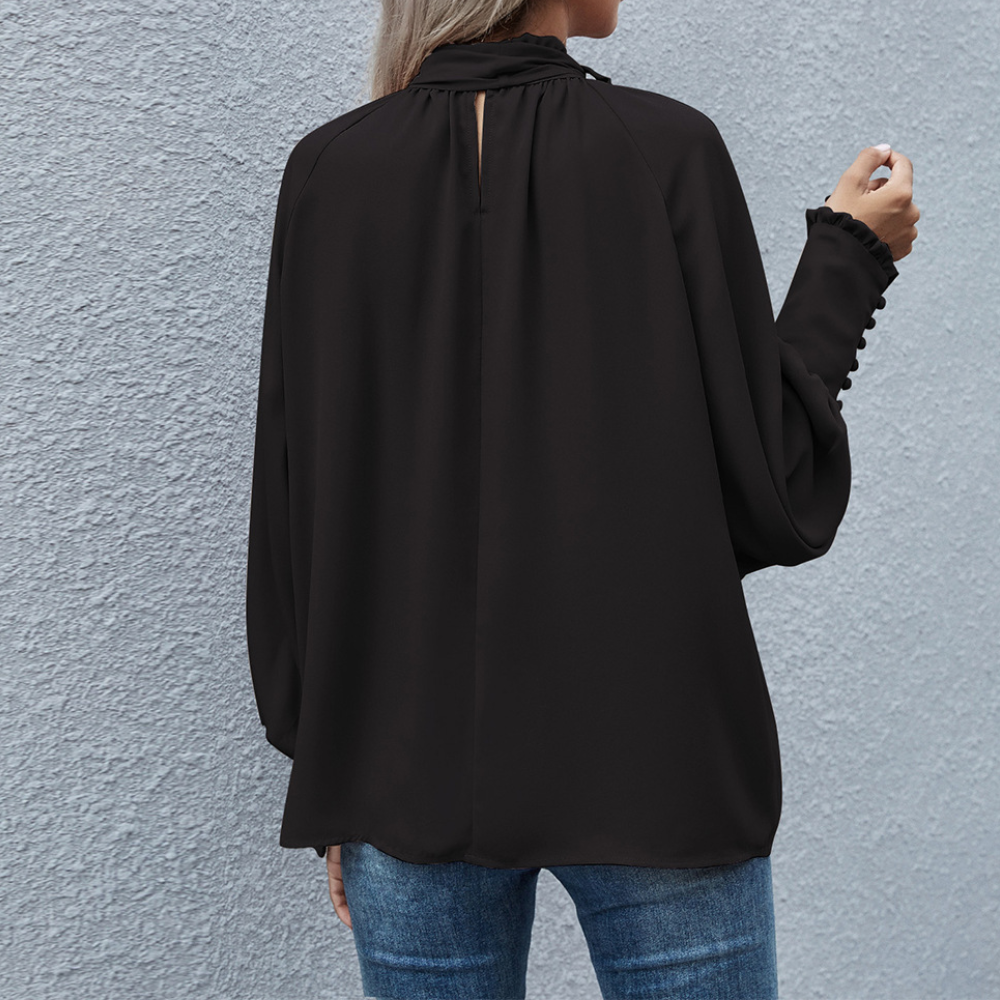 Frau in schwarzer Bluse mit langen Ärmeln und Jeans vor grauer Wand.