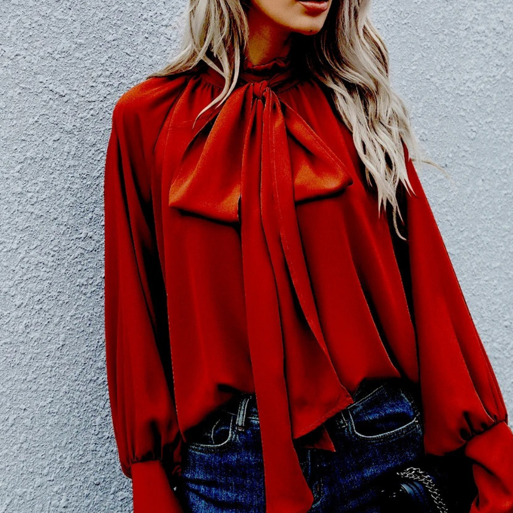 Frau in roter Seidenbluse mit Schleife, blonde Haare, lässiger Stil, vor grauer Wand.