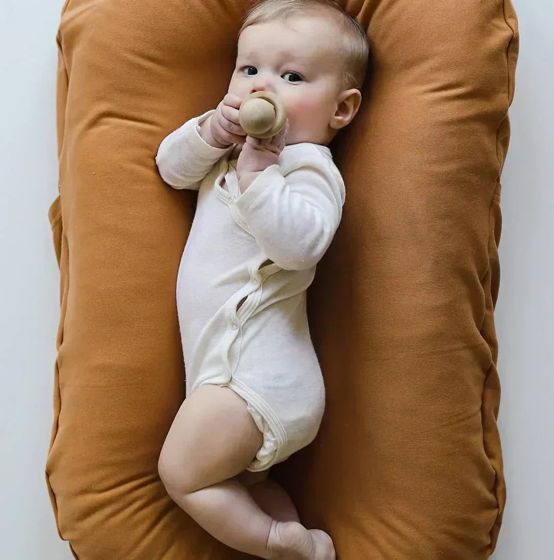 Baby in a cream bodysuit chews a wooden teether in a caramel-colored lounger 4