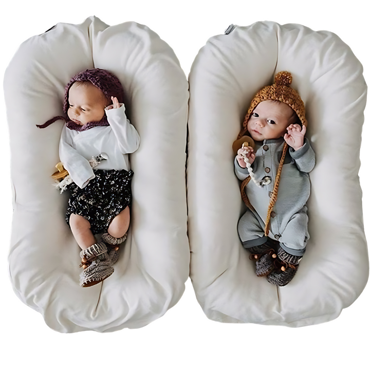 Two newborns in knit hats and outfits lie in two side-by-side cream nest cushions 5