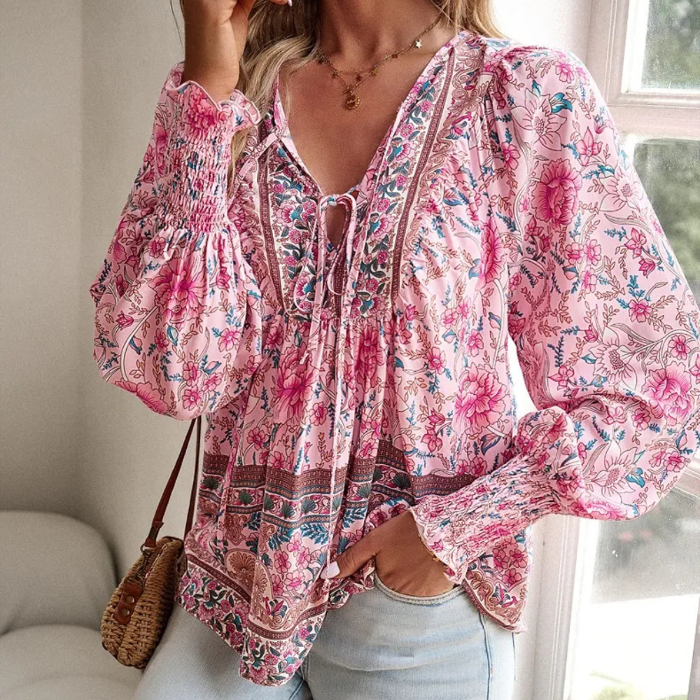 Frau in rosa geblümter Bluse mit langen Ärmeln, Boho-Stil, vor einem Fenster stehend.