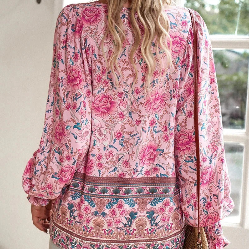 Frau in rosa geblümter Bluse, Boho-Stil, langärmelig, vor einem Fenster stehend.