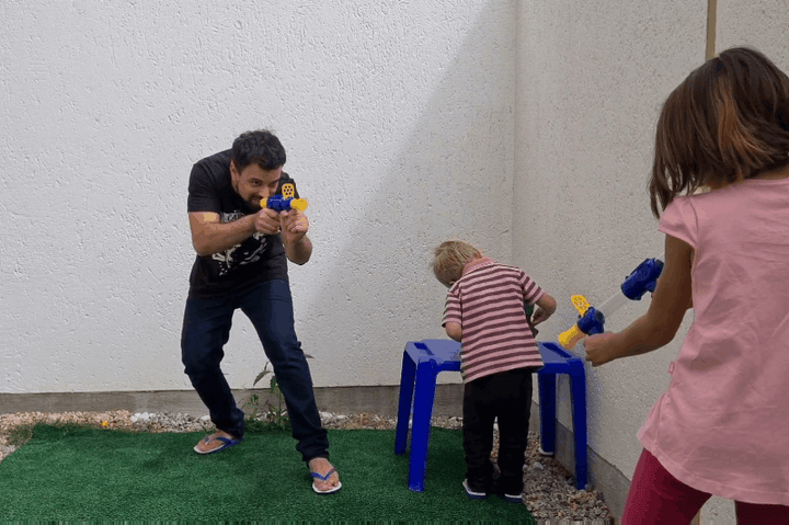 Man and a girl aim blue and yellow blasters outdoors while a toddler stands at a blue stool 10