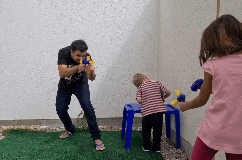 Man, small child and girl play outdoors in a walled yard, aiming blue and yellow foam blasters at each other 10