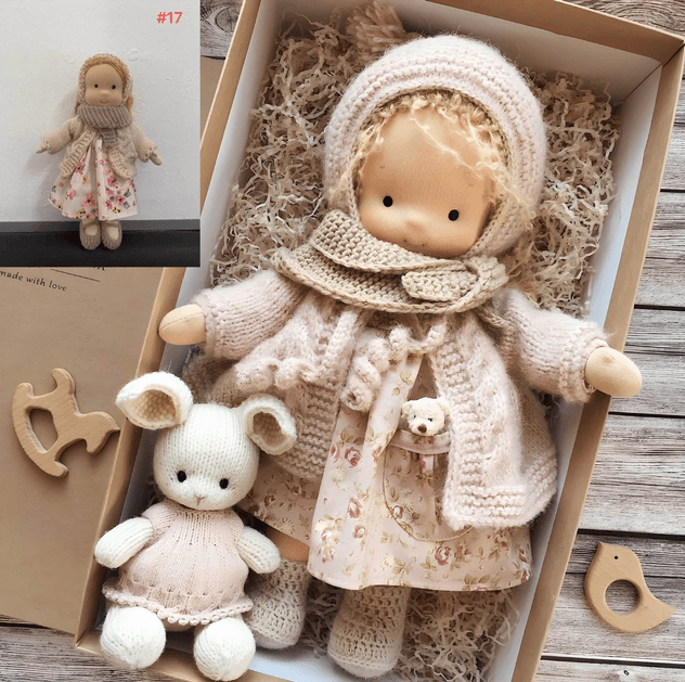 Doll in cream knit hood, fuzzy cardigan and floral dress rests in gift box beside a white knitted bunny 3