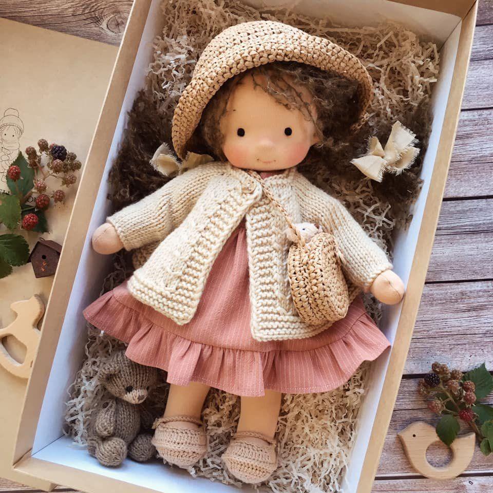 Doll with dark curly hair in woven straw hat, cream cardigan and rose ruffled dress, with a small knit bear 7