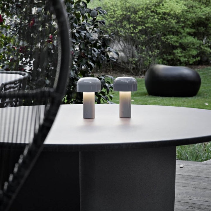 Pair of small gray mushroom lamps lit on a round outdoor table with a garden behind 22