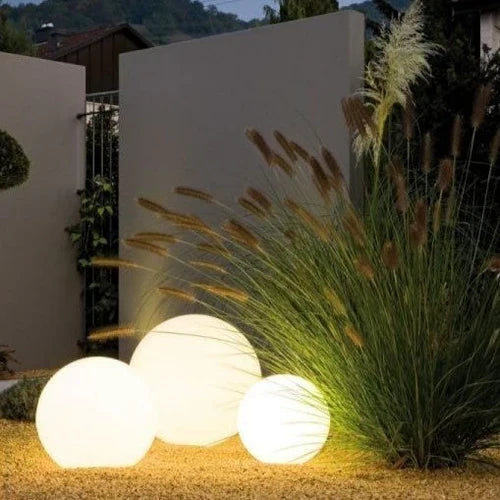 Three white glowing spheres on gravel beside tall ornamental grass and a garden wall 10