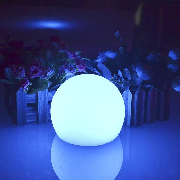 Small sphere glowing blue on a reflective surface in front of a miniature fence and flowers 11