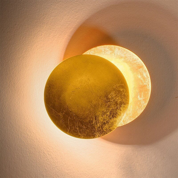 Gold wall lamp with two overlapping discs, the rear one softly glowing against a textured pale wall 12