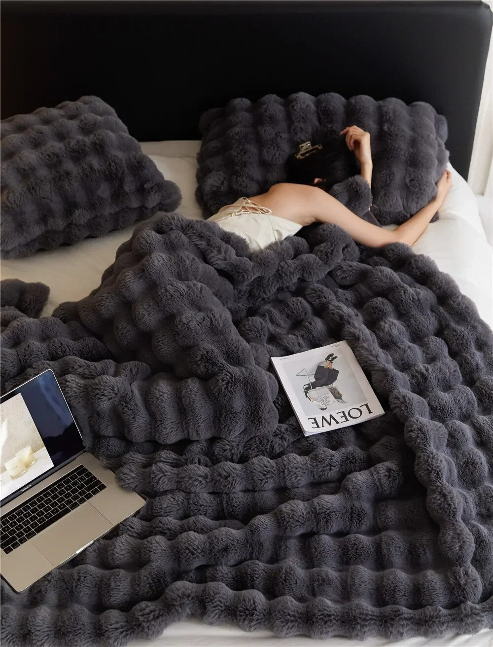 Woman lying under a dark gray plush blanket with matching pillows, a laptop and a magazine beside her 3
