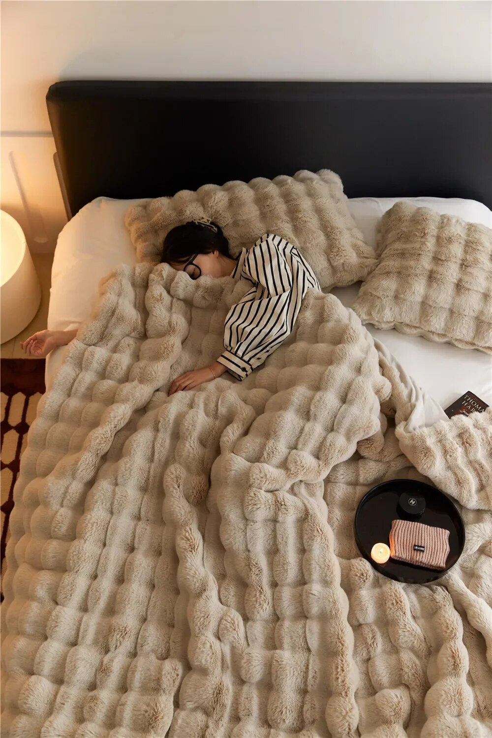 Person sleeping in bed under a beige bubble-textured plush throw, with matching pillows and a candle tray 2